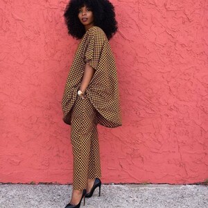 May include: A woman wearing a black and gold patterned trouser suit with a loose-fitting top and long trousers. She is standing in front of a pink wall and wearing black high heels.