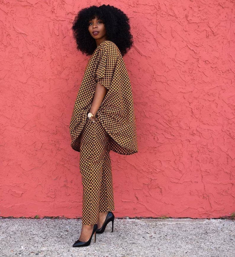 May include: A woman wearing a black and gold patterned trouser suit with a loose-fitting top and long trousers. She is standing in front of a pink wall and wearing black high heels.