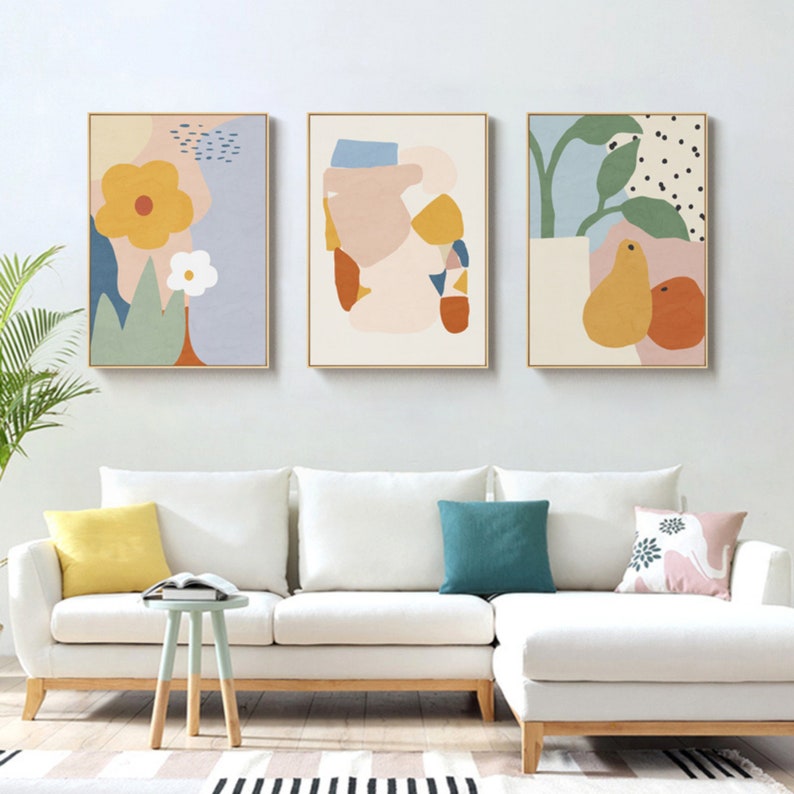May include: Three framed abstract art prints. The artwork features colorful shapes and floral designs in shades of yellow, blue, orange, and pink. The prints are displayed above a white sofa with colorful throw pillows.
