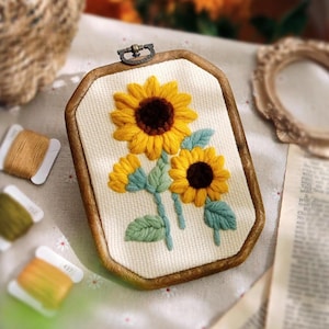 May include: Embroidered sunflower art piece in a wooden frame. The sunflowers are yellow with brown centers and green leaves. The frame has a metal clasp at the top. Spools of thread in various colors are in the background.