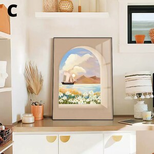 May include: A framed print of a seascape with a sailboat in the distance, framed by an archway with white flowers in the foreground. The print is in a black frame with a beige mat.