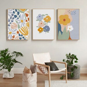 May include: Three framed floral art prints. The artwork features colorful, abstract flower designs in shades of yellow, blue, orange, and pink. The frames are gold. The art is displayed above a chair and plants.