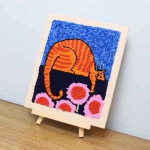 May include: A framed textile art piece featuring a stylized orange tabby cat with a curved back, set against a blue sky and dark blue ground with pink and orange flowers. The artwork is displayed on a small wooden easel.