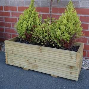May include: A wooden planter box filled with green plants. The box is made of light-coloured wood and sits on small wooden blocks. The plants include a topiary-style tree and several smaller, cone-shaped shrubs, all with vibrant green foliage.