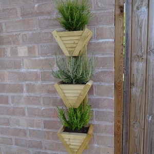 May include: A wall-mounted wooden planter with three triangular sections, each filled with green plants. The planter is fixed to a red brick wall. The natural wood finish contrasts with the green foliage, creating a decorative display.