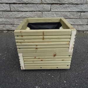 May include: A square wooden planter box with a light green finish. The sides are constructed from horizontal wooden slats, and the top edge has a square frame. The interior contains a black liner. Suitable for outdoor gardening.