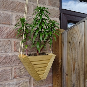 May include: A wooden, triangular hanging planter filled with green plants. The planter is suspended by twine and mounted on a brick wall. The planter has a slatted design on the front.