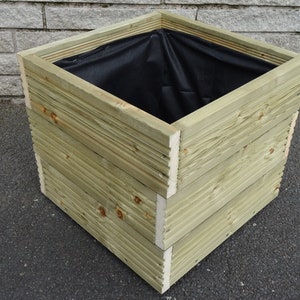 May include: A square wooden planter, constructed from horizontal wooden planks. The natural wood has a light green hue. The interior is lined with black fabric. The planter is suitable for gardens and patios, and is approximately 40cm x 40cm.
