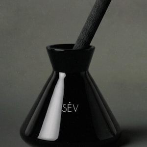 May include: A glossy black ceramic diffuser vessel with a wide, conical base and a narrow neck. The word SÈV is printed in white on the front. A single, textured, dark charcoal-coloured wooden reed stick is inserted into the opening of the vessel.