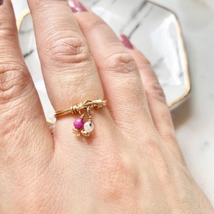 May include: A gold wire ring with two dangling charms. The charms are small, round beads, one pink and one white.