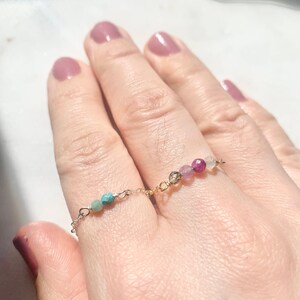 Op de afbeelding: Twee delicate gouden ringen met gekleurde edelsteen kralen. De bovenste ring heeft roze, paarse en rode kralen, terwijl de onderste ring turquoise kralen heeft.
