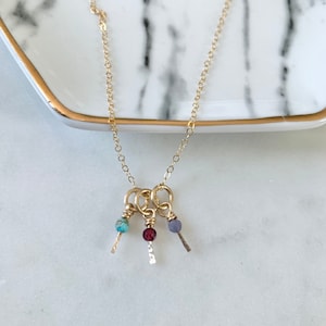 Puede incluir: Un collar de cadena de oro con tres pequeños colgantes. Cada colgante tiene una piedra preciosa de color diferente: turquesa, roja y morada.