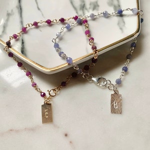 Puede incluir: Dos pulseras de cuentas con piedras preciosas rosas y moradas. Una pulsera tiene un cierre dorado y un dije rectangular con la letra "E". La otra pulsera tiene un cierre plateado y un dije con la letra "B".