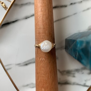 Könnte beinhalten: Ein Ring aus goldfarbenem Draht mit einer weißen Perle. Der Ring wird auf einer Holzoberfläche gezeigt.
