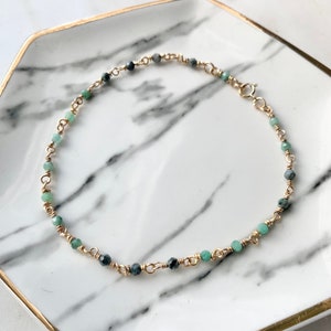 Könnte beinhalten: Ein zartes Armband aus einer goldenen Kette mit abwechselnd grünen und grauen Edelsteinperlen.