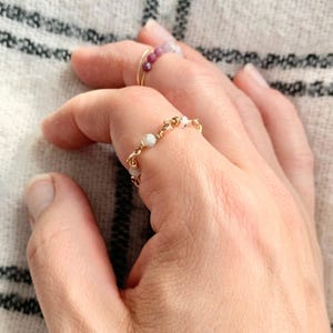 Könnte beinhalten: Nahaufnahme von zwei goldenen Ringen an einer Hand. Ein Ring hat kleine, runde, weiße Perlen, die mit Golddraht aufgefädelt sind. Der andere Ring hat kleine, runde, rote Perlen. Die Ringe befinden sich an einer Hand mit neutralem Hintergrund.