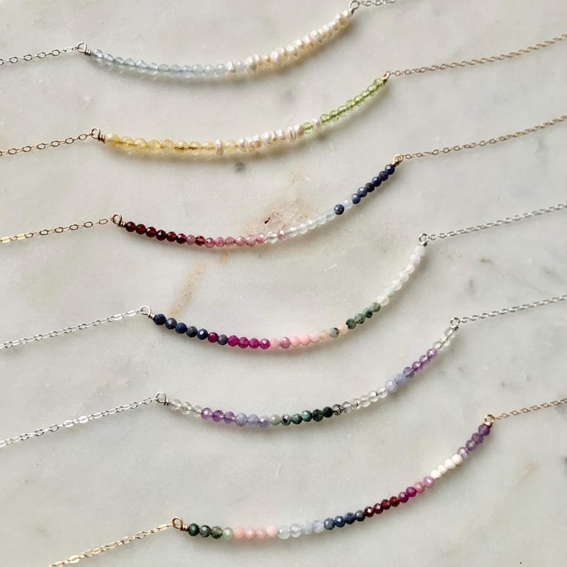 May include: Six delicate necklaces with graduated gemstone beads in various colors, including blue, green, pink, and red. The necklaces are arranged in a staggered pattern on a white marble surface.