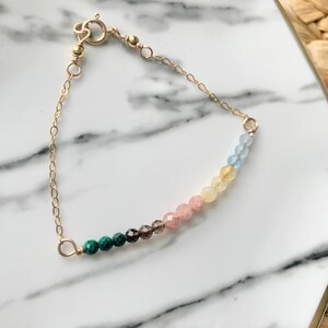 May include: A gold chain bracelet with a rainbow of gemstone beads. The beads are green, brown, pink, yellow, and blue.