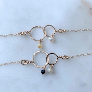 Puede incluir: Dos collares de oro con cadenas delicadas y círculos entrelazados. Cada collar presenta una pequeña perla y un colgante de piedra preciosa. Un colgante es una piedra negra, el otro es una piedra amarilla.