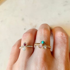 以下が含まれることがあります： 宝石のアクセントが付いた、金色のワイヤーで巻かれた2つのリング。左側のリングは、白いムーンストーンと赤い宝石が特徴です。右側のリングは、ターコイズの宝石、ピンクの宝石、白いムーンストーンが特徴です。