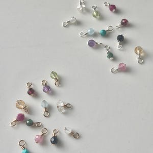 May include: A collection of gemstone charms in various colors, including pink, green, purple, and clear. Each charm features a small, round gemstone attached to a gold-colored wire loop. The charms are scattered on a white surface.
