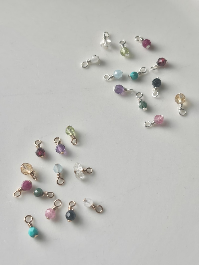 May include: A collection of gemstone charms in various colors, including pink, green, purple, and clear. Each charm features a small, round gemstone attached to a gold-colored wire loop. The charms are scattered on a white surface.