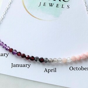 May include: A silver chain necklace with a row of gemstone beads in a gradient of colors, starting with purple and ending with pink. The beads are labeled with the months of the year, starting with February and ending with October. The necklace is on a white card with the words "JEWELS" printed on it.