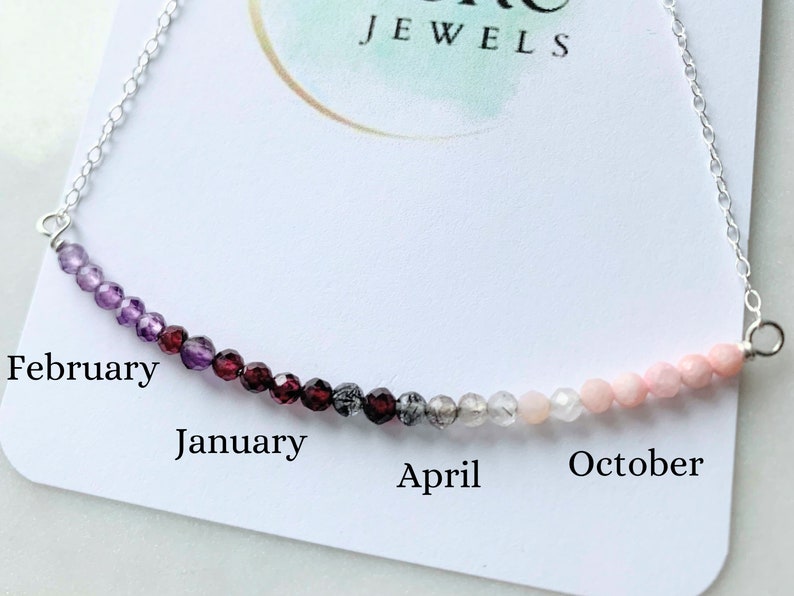 May include: A silver chain necklace with a row of gemstone beads in a gradient of colors, starting with purple and ending with pink. The beads are labeled with the months of the year, starting with February and ending with October. The necklace is on a white card with the words "JEWELS" printed on it.