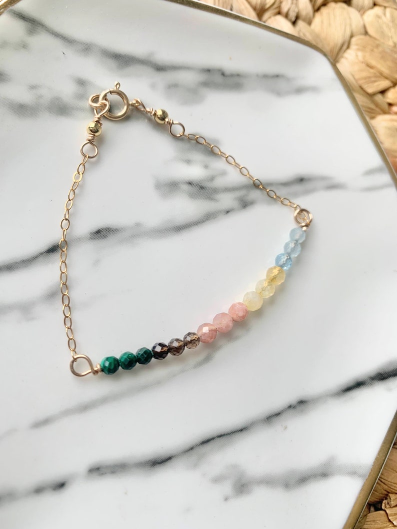 May include: A gold chain bracelet with a rainbow of gemstone beads. The beads are green, brown, pink, yellow, and blue.