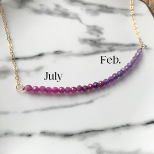Pode incluir: Um colar de corrente de ouro com uma fileira de pedras preciosas rosa e roxas. As pedras preciosas são dispostas em degradê, com rosa à esquerda e roxo à direita. O colar é etiquetado com "Julho" à esquerda e "Fev." à direita.