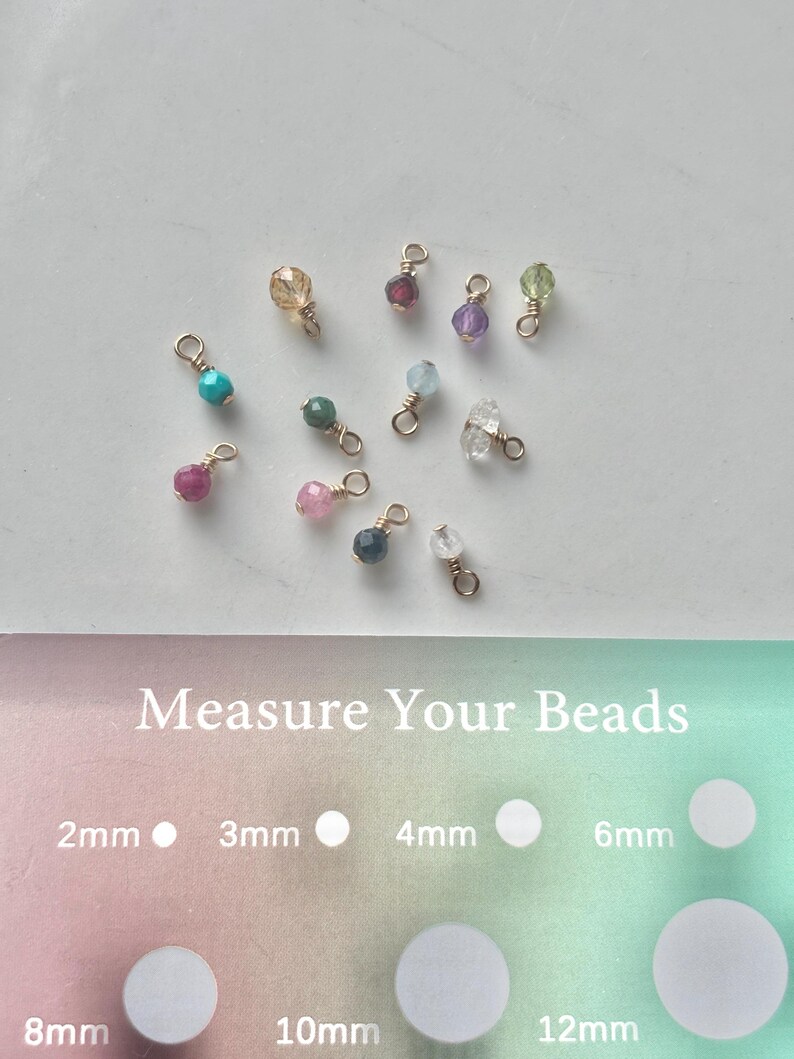 May include: A collection of colorful gemstone beads with gold-colored findings. The beads vary in color, including teal, pink, green, and purple. The image also includes a guide to measure the beads, with sizes ranging from 2mm to 12mm.