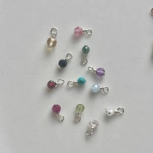 May include: A collection of small, colorful gemstone charms with silver-colored wire loops. The charms feature various faceted beads in shades of pink, green, purple, blue, and clear, perfect for jewelry making or crafting.