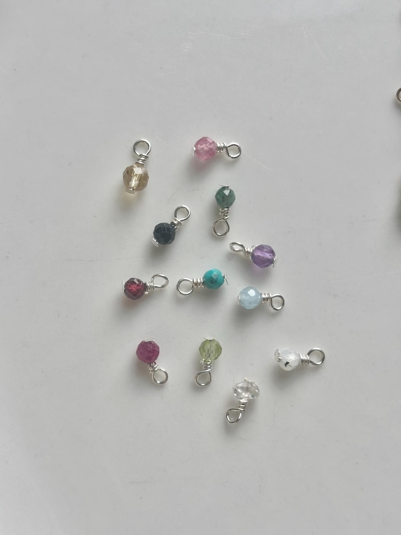 May include: A collection of small, colorful gemstone charms with silver-colored wire loops. The charms feature various faceted beads in shades of pink, green, purple, blue, and clear, perfect for jewelry making or crafting.