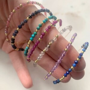 May include: A collection of delicate beaded bracelets in various colors, including pink, blue, green, and purple. Each bracelet features small, faceted gemstones and gold-colored beads. The bracelets are displayed on a light background.
