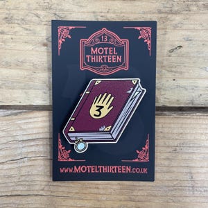 May include: A red and gold enamel pin featuring a closed book with a hand and the number 3 on the cover. The pin is on a black background with the text "Motel Thirteen" and "www.motelthirteen.co.uk".