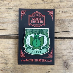 May include: Green and white enamel pin featuring a green plant with a face and the text "Hexside Plant" in a white circle with white stars. The pin is on a black background with the text "Motel Thirteen" in red.