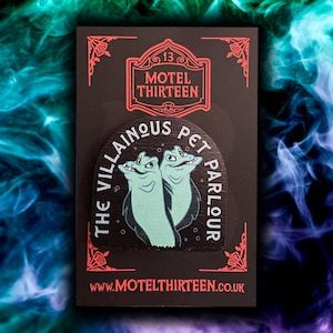 May include: A black pin featuring the text "THE VILLAINOUS PET PARLOUR" and two cartoonish, light teal creatures. The pin also has the text "MOTEL THIRTEEN" and "www.MOTELTHIRTEEN.co.uk" in red.