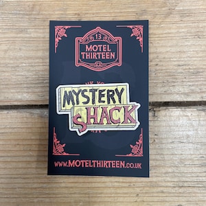 May include: A black card featuring a red and white "MOTEL THIRTEEN" logo and a "MYSTERY SHACK" pin. The pin has a yellow background with red and black text. The website address www.MOTELTHIRTEEN.co.uk is at the bottom.