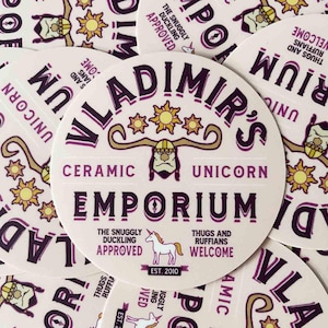 May include: A pink and purple round sticker with the text "Vladimir's Ceramic Unicorn Emporium" in a stylised font. The sticker features a unicorn, a viking with horns, and the text "The Snuggly Duckling Approved" and "Thugs and Ruffians Welcome".
