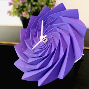 Peut inclure: Horloge en origami violette avec des aiguilles blanches. L'horloge a la forme d'une spirale.