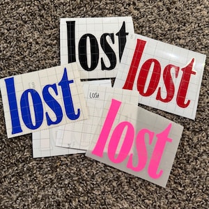 May include: A collection of vinyl stickers with the word "lost" in various colors: black, red, blue, and pink. Each sticker is rectangular and has a grid backing. The stickers are arranged on a brown, textured surface.