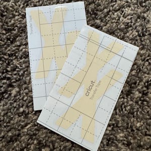 Puede incluir: Dos hojas de cinta de transferencia Cricut con un patrón de cuadrícula y un diseño amarillo claro. El texto "Transfer Tape" está impreso verticalmente en el lado izquierdo de las hojas. El logotipo de Cricut es visible en el lado derecho.