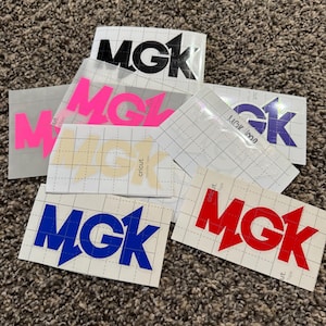 Puede incluir: Una colección de pegatinas de vinilo con las letras "MGK" en varios colores, incluyendo negro, rosa, azul y rojo. Las pegatinas están sobre un respaldo blanco con un patrón de cuadrícula.