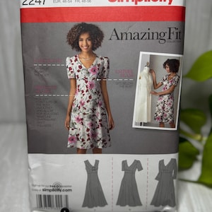 Può includere: Una busta di cartamodello Simplicity 2247 per un abito in taglia BB 20W-28W. Il fronte mostra un modello che indossa un abito bianco con stampa floreale rosa e grigia, maniche corte e scollo a V. Include diagrammi di tre varianti di abito.