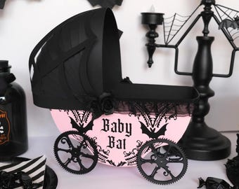 May include: A pink and black gothic-themed baby carriage with the words "Baby Bat" on the side. The carriage has black wheels, a black hood, and is decorated with bats and roses. A black candelabra and other gothic decorations are in the background.