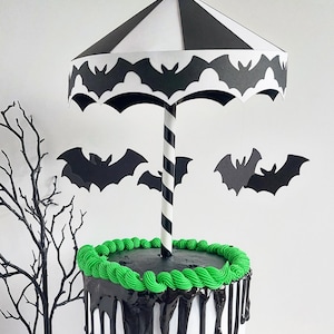 May include: A two-tiered cake decorated with black and green frosting. The top tier has a black and white striped paper umbrella with bats around the edge. The umbrella is attached to a black and white striped straw.