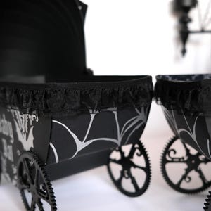 May include: Black and white decorative carriage with a black lace trim. The carriage has black wheels and a patterned design with bats and spiderwebs. The carriage is a Halloween or gothic-themed item.