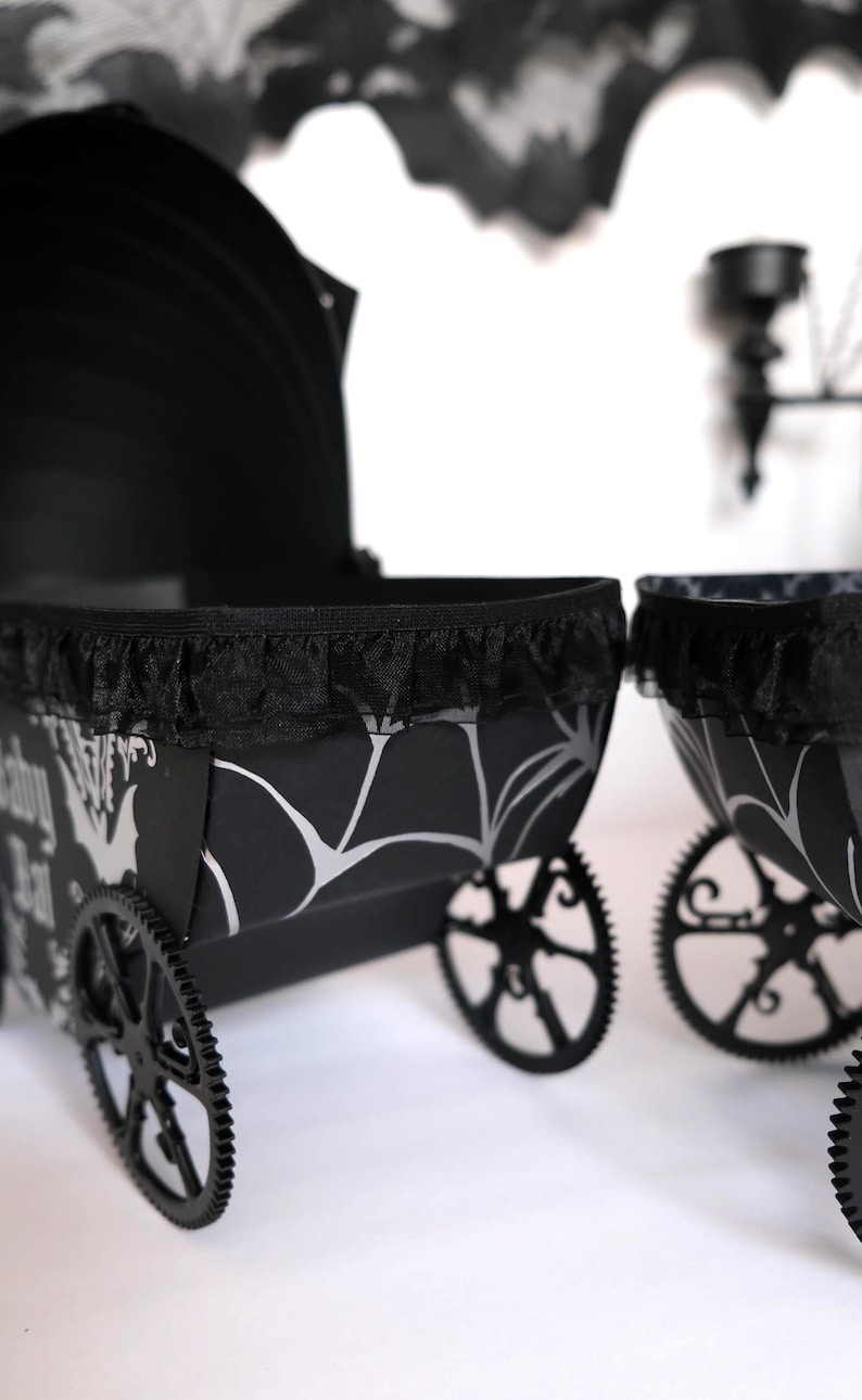 May include: Black and white decorative carriage with a black lace trim. The carriage has black wheels and a patterned design with bats and spiderwebs. The carriage is a Halloween or gothic-themed item.