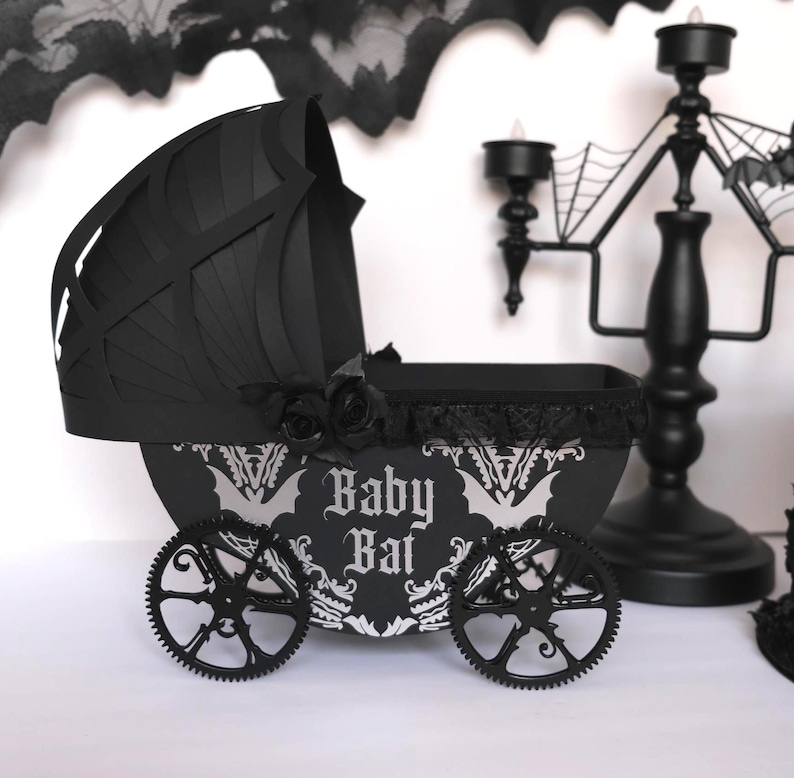 May include: A black gothic-themed baby carriage. The carriage has a bat-shaped hood, decorative wheels, and the words "Baby Bat" in a stylized font. Black roses and lace trim enhance the spooky design. A candelabra is visible in the background.