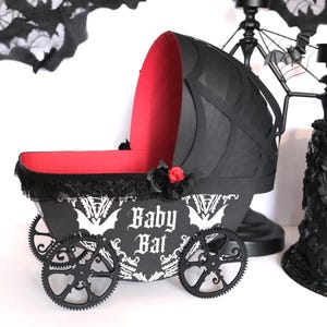 May include: A decorative black and red baby carriage featuring the words "Baby Bat" in white gothic script. The carriage has gear-shaped wheels and is decorated with black lace and a red rose. The carriage is approximately 30 cm long.